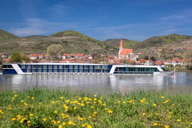 Modern river cruise ship with glass windows sails along the Danube River in front of the village of Weissenkirchen, Austria, with a church steeple and vineyards behind.