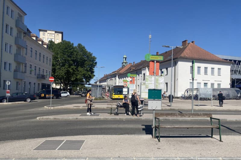 People waiting at a bus stop near Krems an der Donau railway station in Austria, with a yellow VOR bus approaching and old buildings in the background.