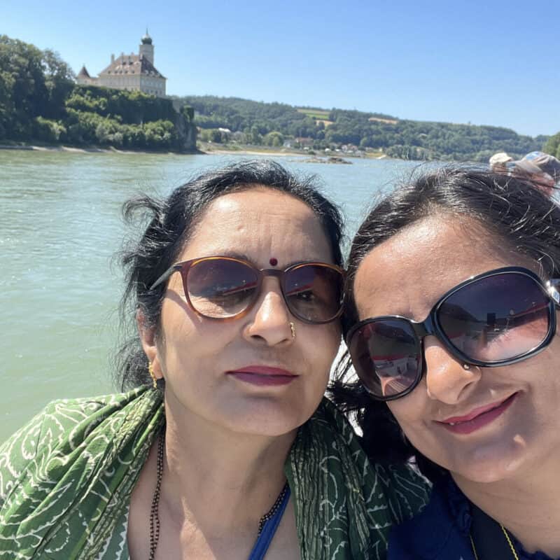 Two women smiling in a selfie on a sunny day, with the Danube River and Schönbühel Castle in the background in Wachau Valley, Austria.