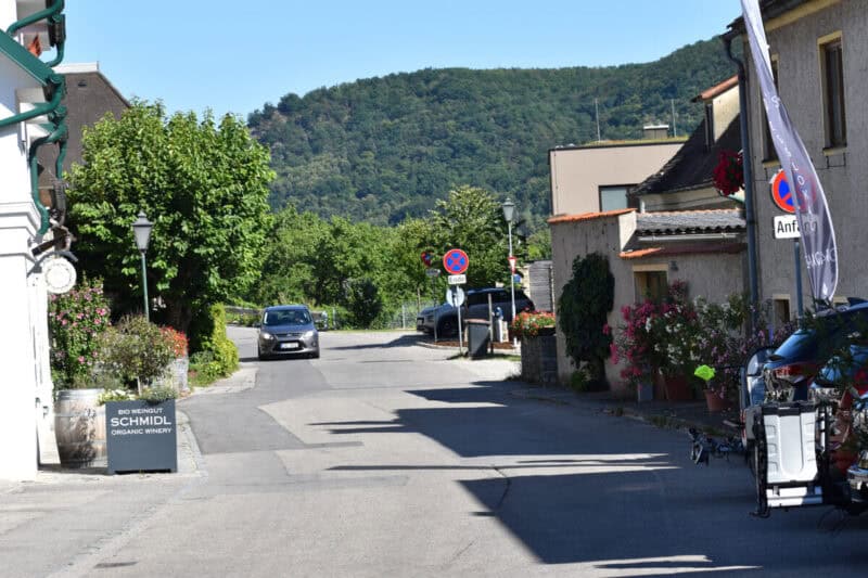 Street scene in a small Austrian village in Wachau Valley, with cars parked by quaint houses, flowering plants, and green hills rising in the background.