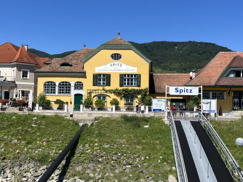 Bright yellow building marked “Spitz” with green shutters and white trim sits beside the Danube River, with a ramp leading down to a boat landing, under sunny blue skies.