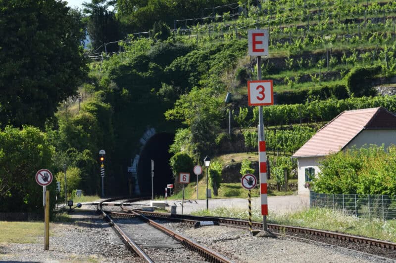 Railway tracks leading into a tunnel through green vineyards on a sunny day in Wachau Valley, Austria, with signs and signal lights along the tracks.