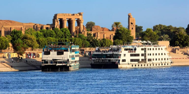 Two large cruise ships docked along the Nile River in front of the ruins of Kom Ombo Temple in Egypt, under a bright blue sky