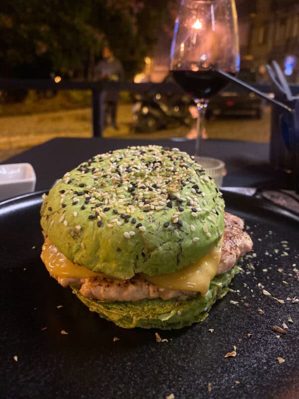 Green bun veg burger with melted cheese and seeds served on a black plate at an outdoor restaurant in Portugal.