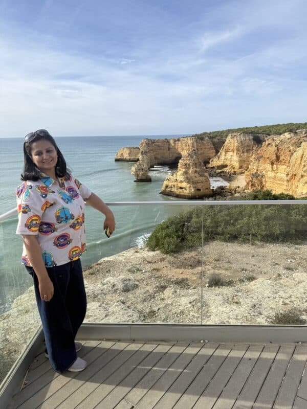 Woman standing at a glass viewpoint with Algarve sea stacks and cliffs in Praia da Marinha, Portugal, with calm ocean and clear sky in the background.