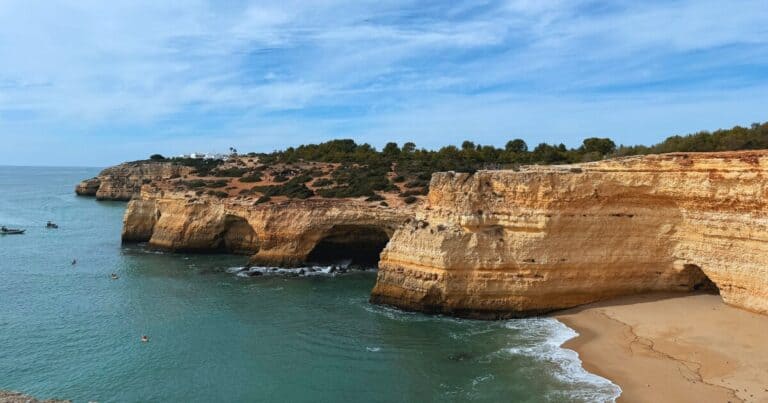Wide view of golden Algarve cliffs and sea caves along the Portugal coast with calm blue water and sandy beach below