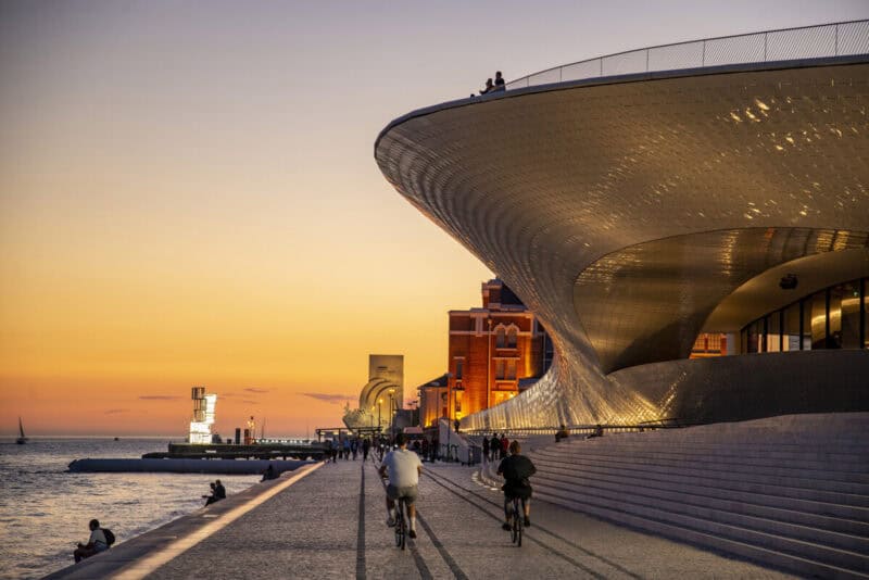 Avenida Brasília in Lisbon with cyclists passing MAAT at sunset along the Tagus waterfront
