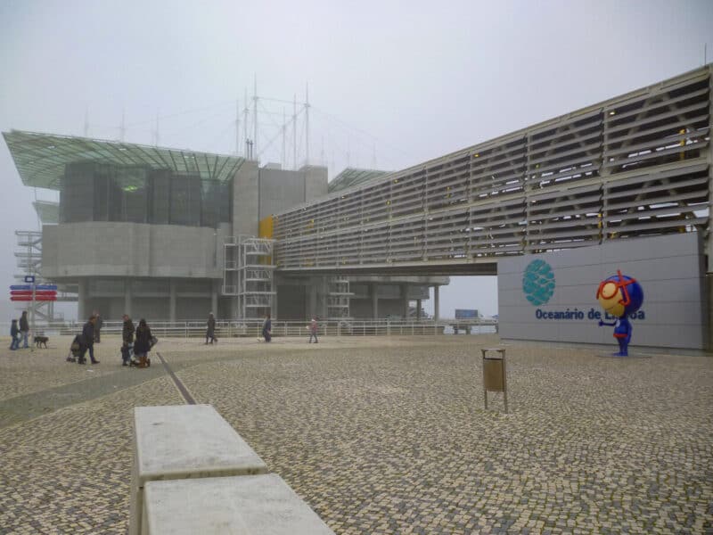 Oceanário de Lisboa on a foggy and overcast day, with visitors walking across the waterfront plaza near the aquarium entrance.