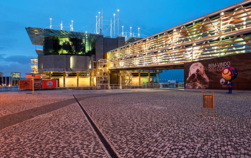 Oceanário de Lisboa illuminated at dusk, with lights reflecting across the modern waterfront building on a calm evening.