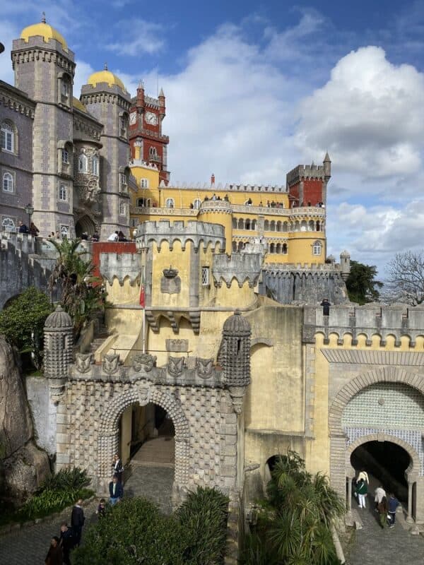 Colourful façades and ornate towers of Pena Palace rising above the entrance arches in Sintra, Portugal, under a dramatic cloudy sky.