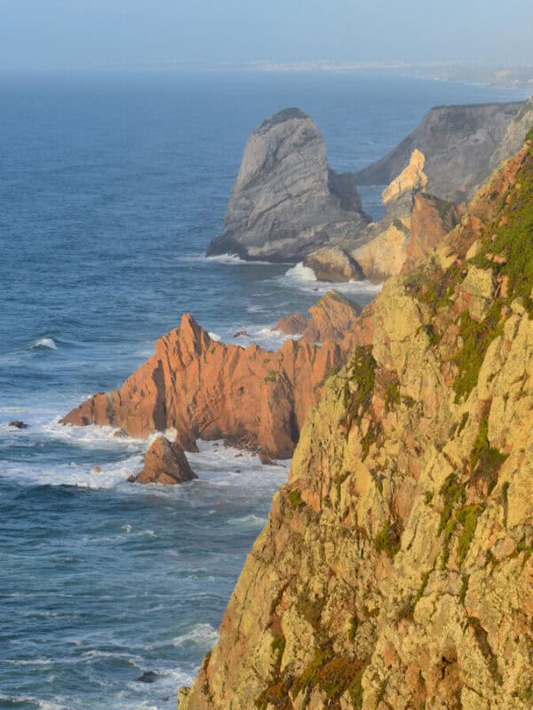 Rugged coastal cliffs and rock formations rise above the Atlantic Ocean, with waves crashing against the shoreline below at Cabo da Roca