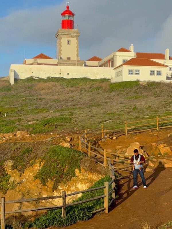 Vacaywork author Ashrith carrying his son while walking downhill at Cabo da Roca with the lighthouse behind