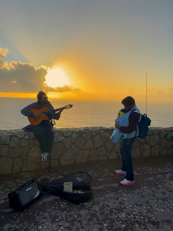 A street musician plays guitar at sunset on a cliff overlooking the sea while vacaywork author ashrith stands nearby holding a child.