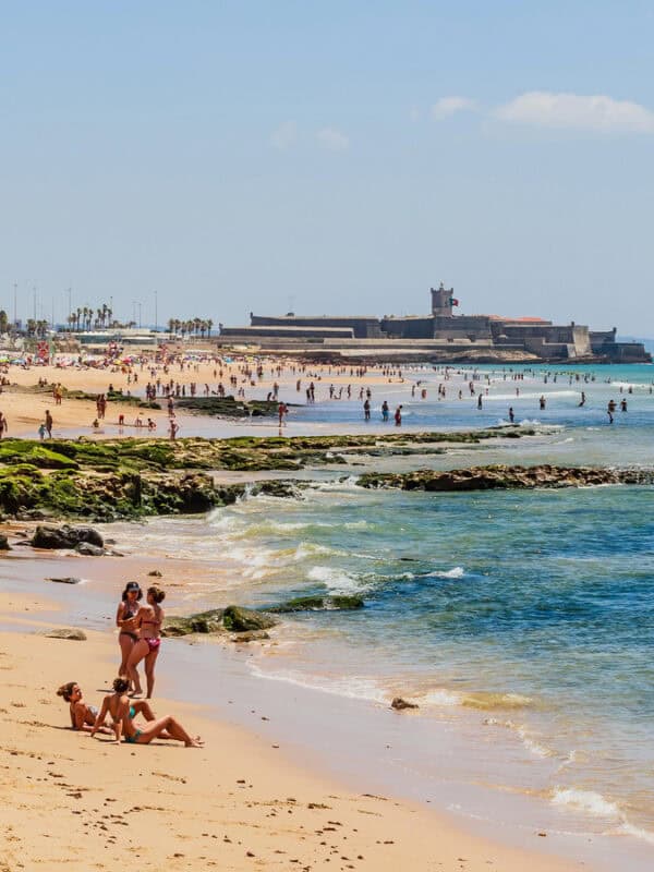 People relax and swim on a sandy beach near a historic seaside fort at carcavelos, with gentle waves and clear blue water.