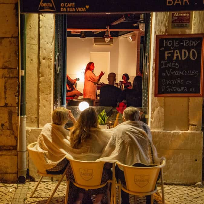 People sit outside a restaurant wrapped in blankets while a Fado singer performs inside for diners under warm evening lights in Lisbon