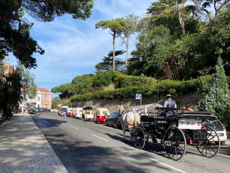 A cobbled foothpath beside asphalt road in Sintra with a horse-drawn carriage, parked tuk-tuks and cars outside the historic centre.