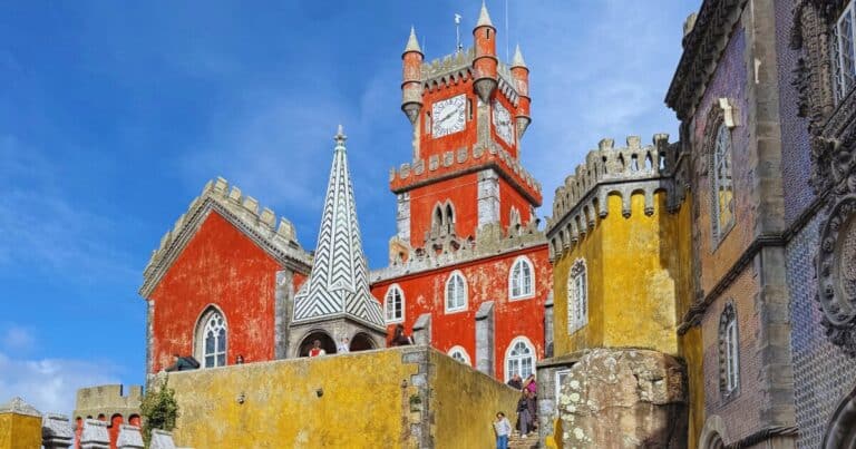 Colourful red clock tower, yellow walls and ornate façades of Pena Palace in Sintra, Portugal, viewed from the courtyard with visitors exploring one of Europe's most famous fairytale castles.