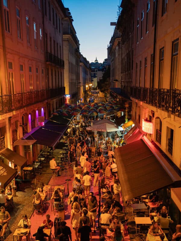 A lively Lisbon street at night filled with outdoor restaurants, colourful umbrellas and people dining and walking between cafés.