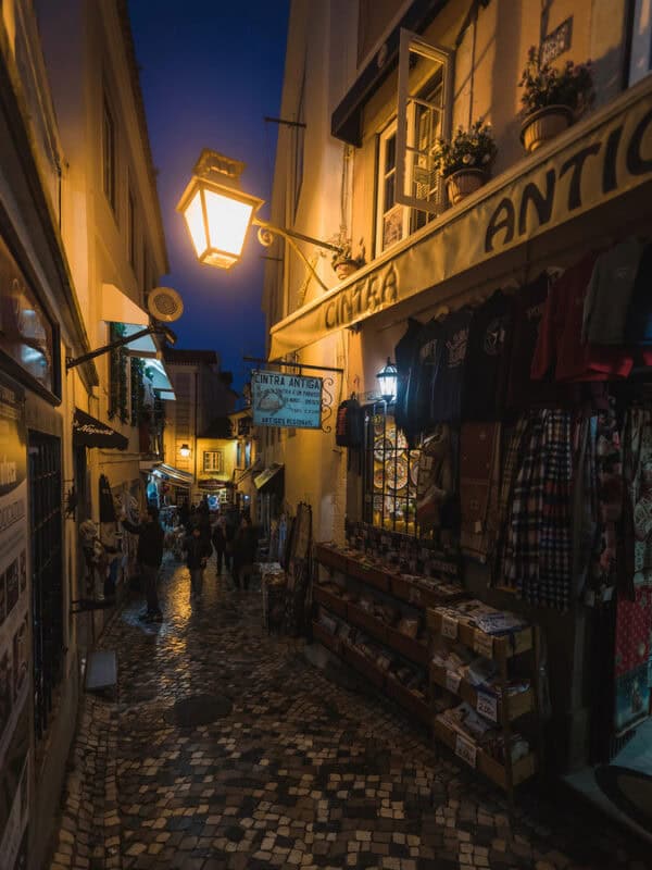 A narrow cobbled street in Sintra lined with closed souvenir shops and old buildings, lit by traditional street lamps after sunset.