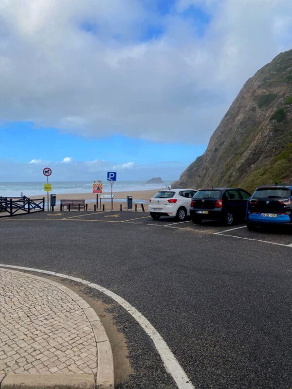Cars parked in the designated parking area near Praia da Adraga, with ocean views from the roadside.