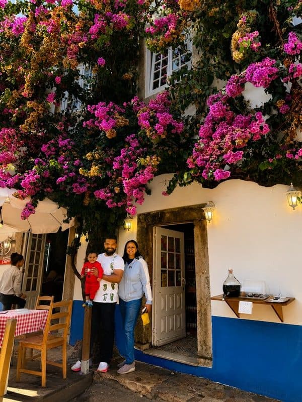 Sahana, Ashrith and their son stand outside Lidador Restaurant in Óbidos, beneath a colourful bougainvillea-covered whitewashed building with blue trim.