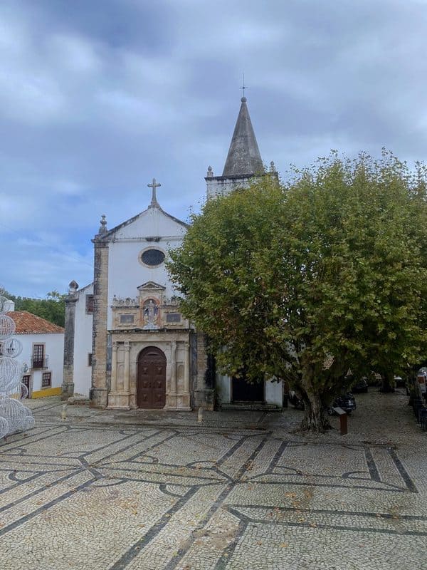 Saint Mary Church in Óbidos with simple Baroque detailing, a tower beside it, patterned cobbled flooring and a large tree in front.