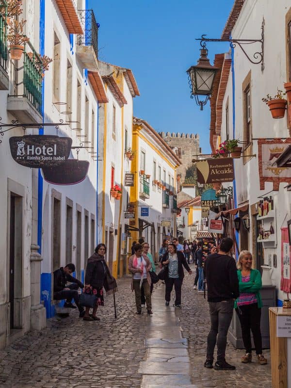 Cobbled Rua Direita in Óbidos lined with whitewashed buildings, colourful shops and Hotel Rainha Santa Isabel, with the castle walls visible at the end of the street.