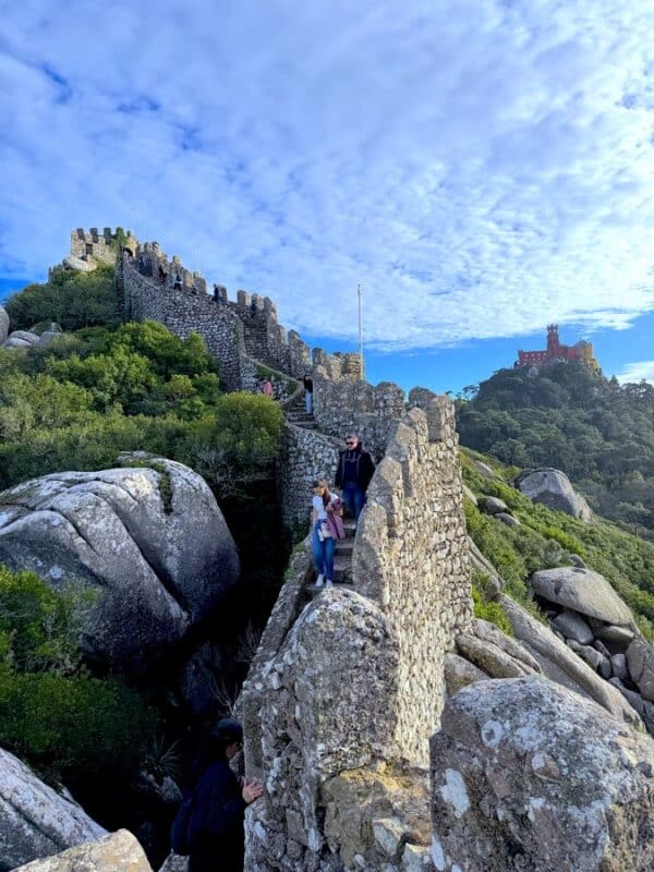 Moorish Castle hike in Sintra with visitors walking along the ancient stone walls and Pena Palace visible across the forested hills.