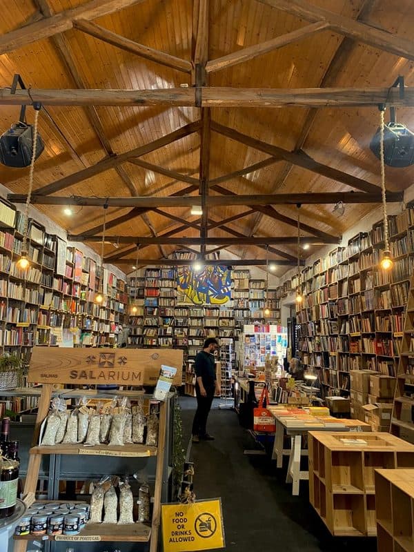Interior of Mercado Biológico de Óbidos, with books lining both walls beneath a wooden vaulted roof, a central display of local products and tables arranged along the shop.