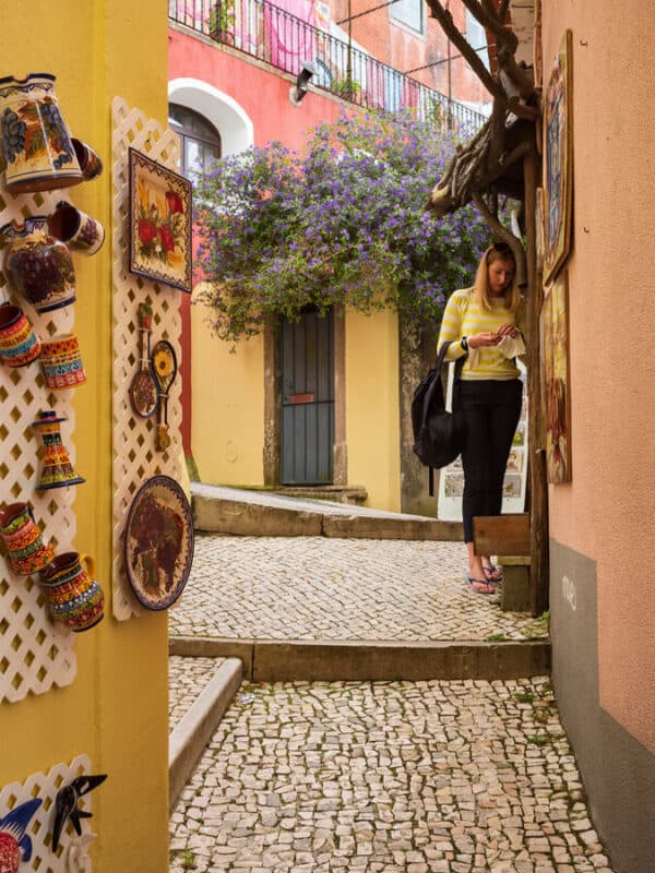 Narrow cobbled lane of Ferreira street in Sintra Portugal with colourful houses, ceramic souvenirs, flowers and a traveller exploring the street.