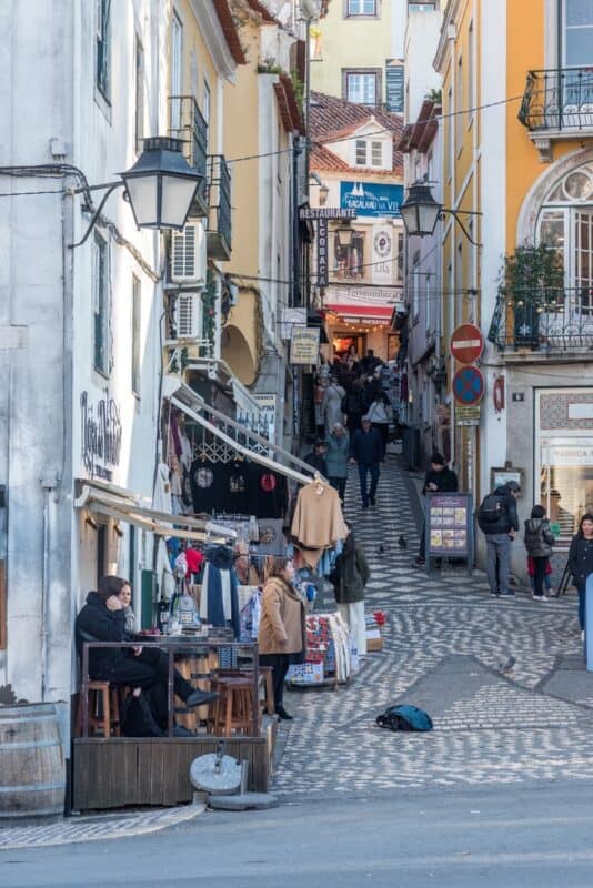 Narrow shopping lane Rua da Ferraria in Sintra seen from the main road, with chequered cobbled stones, historic buildings, shops, cafés and visitors.