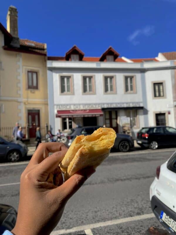 Hand holding a traditional Portuguese pastry on volta do duche street in front of Queijadas da Sapa bakery