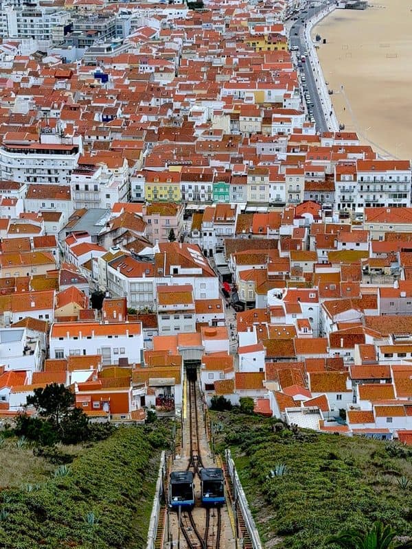 Aerial view of two funicular trains passing each other on the track, with Nazaré town and clusters of red tiled roofs below