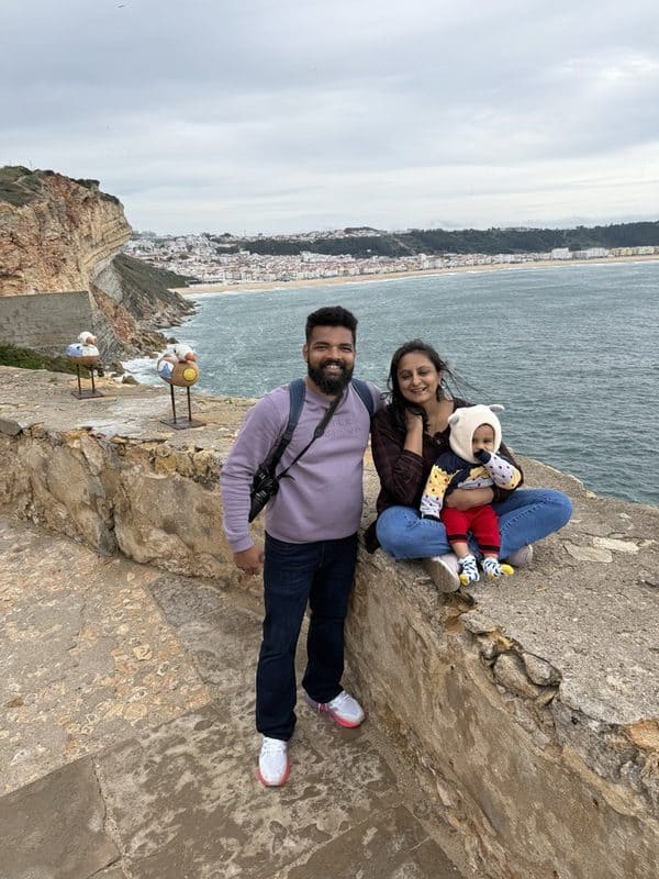 Vacaywork founders Ashrith and Sahana sitting with their son on the lighthouse courtyard wall, with the Atlantic Ocean and Nazaré cliffs behind them
