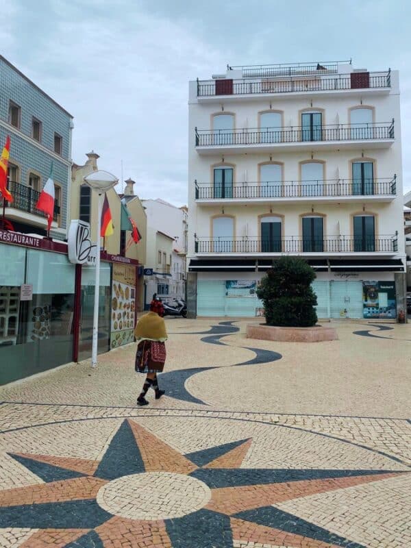 Nazare Portugal square with traditional Portuguese pavement, white buildings and a woman wearing traditional seven layered skirt walking through the centre
