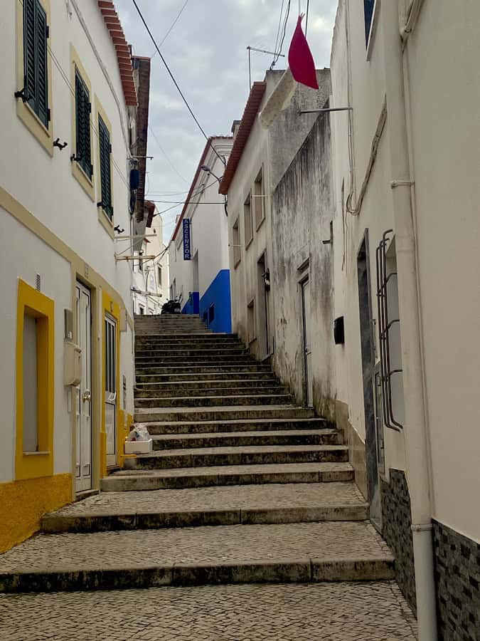 Narrow pedestrian street in Sítio, Nazaré, lined with beige houses and yellow and blue doors, with limited stroller access