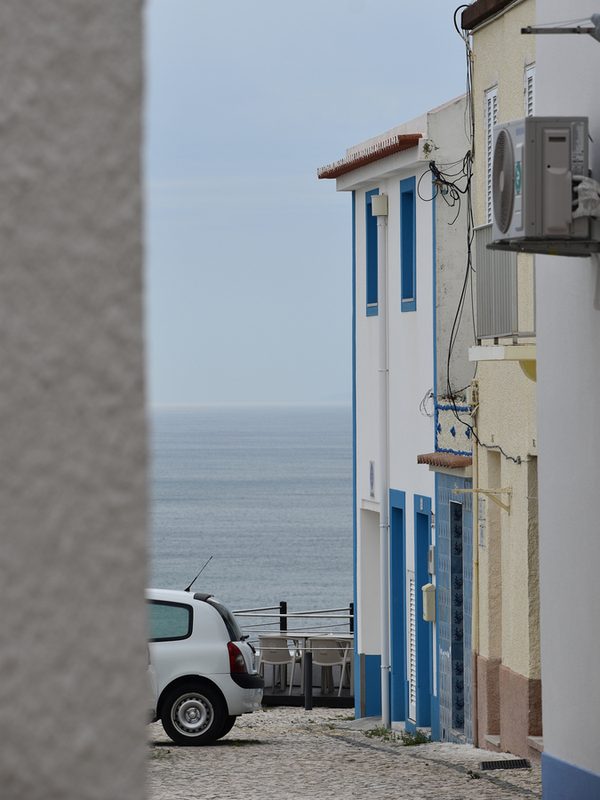 Narrow cobbled streets in Sítio, Nazaré, lined with pastel houses and blue doors, with the street opening towards the Atlantic Ocean